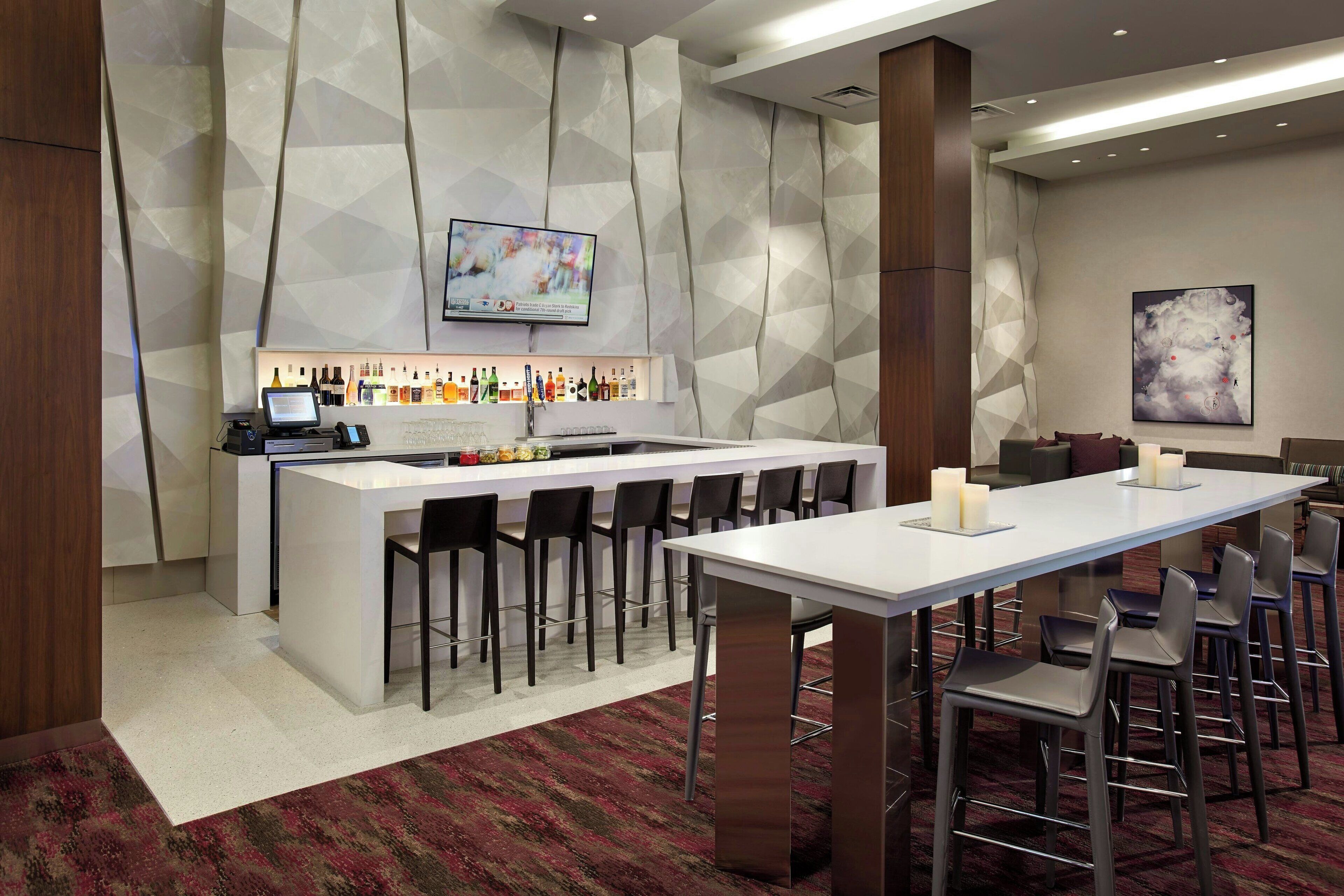 Zona de bar moderna con televisor de pantalla plana, diversos tipos de botellas y una mesa larga con sillas.