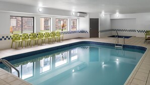 A swimming pool with a tiled deck, a seating area with chairs, and windows with a view of a brick building. - Travelodge by Wyndham Fargo West Acres (Fargo)