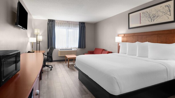 A hotel room with a large bed, a desk with a flat-screen TV, a chair, a small table, and a window with curtains. - Travelodge by Wyndham Fargo West Acres (Fargo)