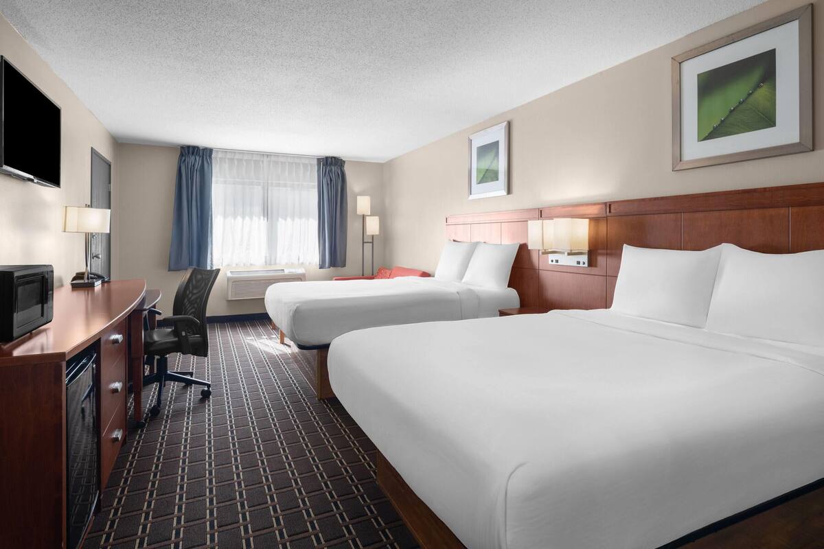 A hotel room with two beds, a desk with a TV, a chair, a window with curtains, and a framed picture on the wall.