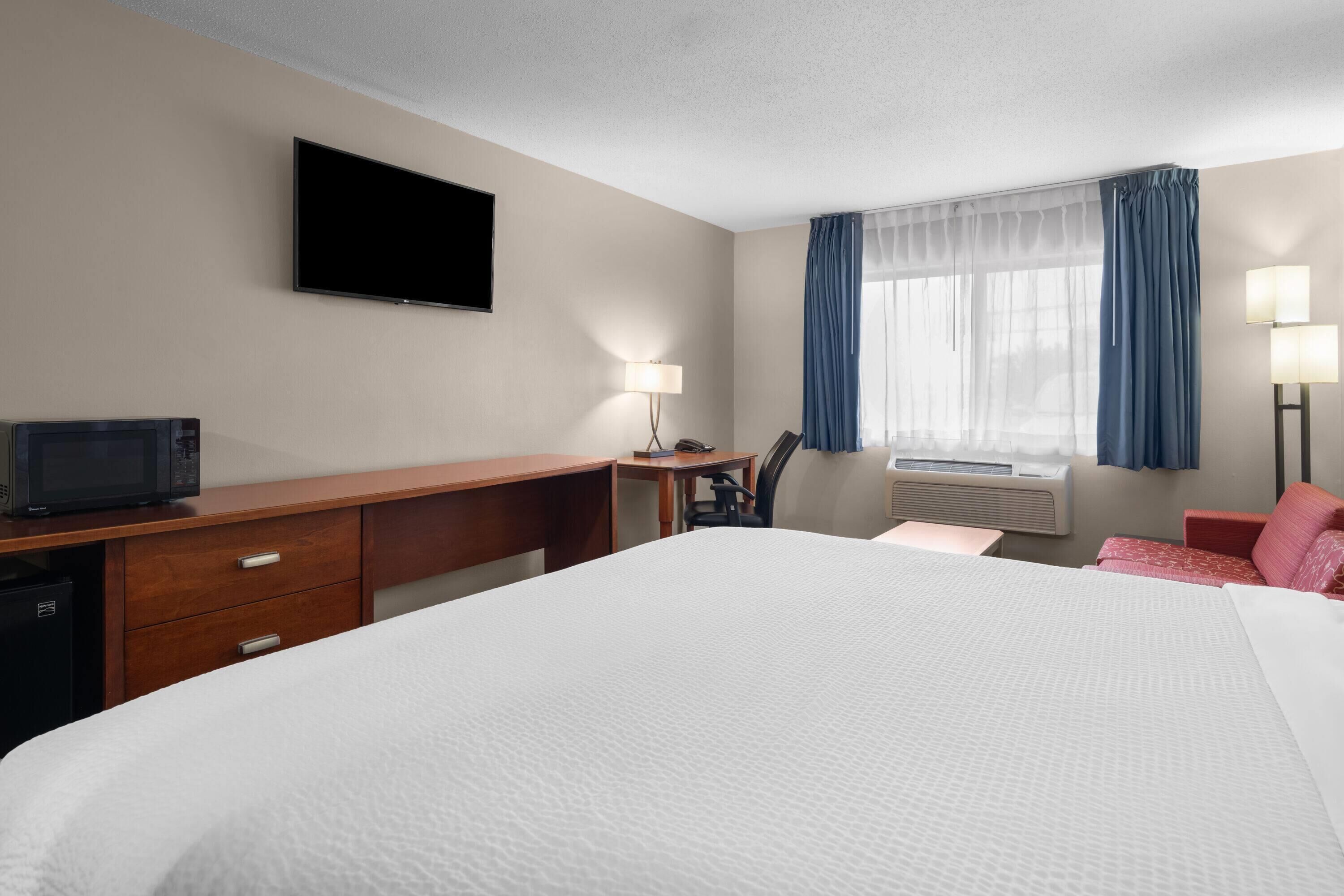 A hotel room with a large bed, a desk with a computer, a television, a lamp, and a window with curtains.