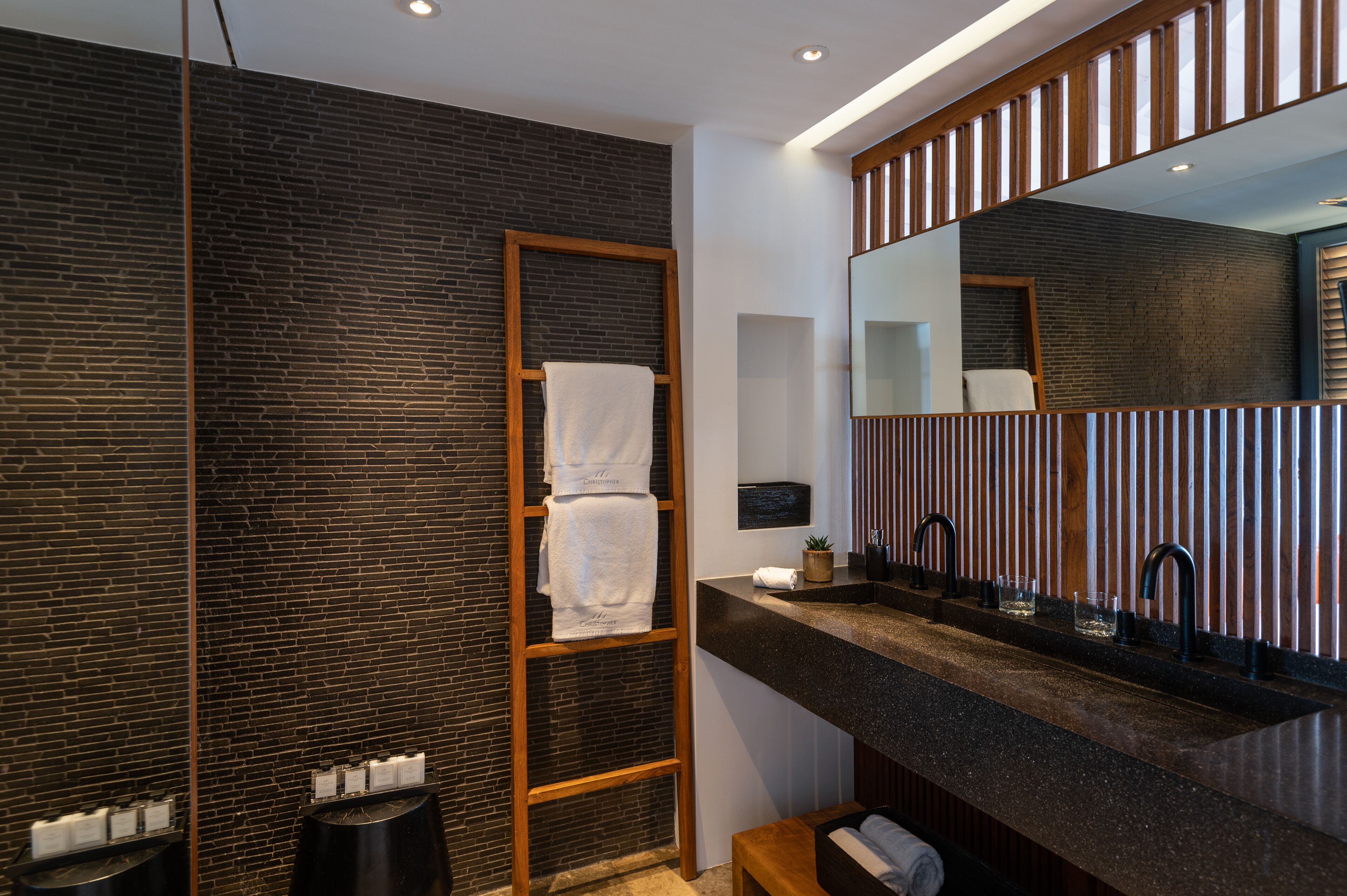 A modern bathroom with a dark brick wall, a wooden towel rack, a large mirror, and a granite countertop with a sink and faucet.