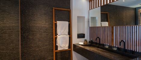 A modern bathroom with a dark brick wall, a wooden towel rack, a large mirror, and a granite countertop with a sink and faucet.