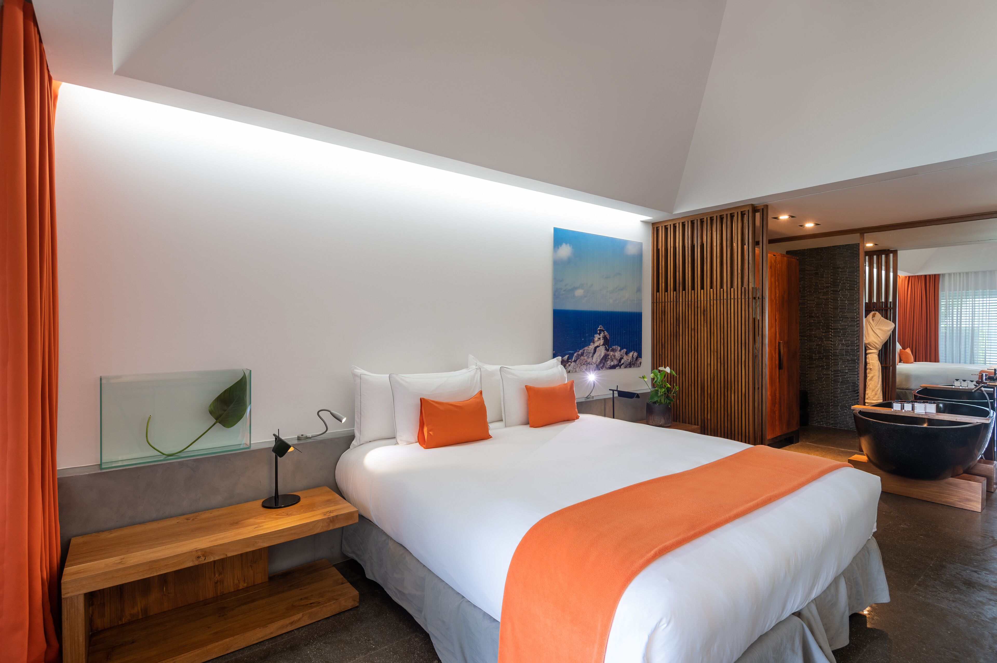 A modern hotel room with a large bed, a wooden bench, a glass table with a leaf, and a stone wall.