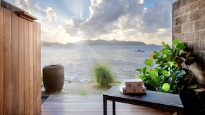 A wooden deck with a view of the sea and mountains, a stone planter, and a table with a towel and a box. - Christopher Hotel (St. Barthelemy)