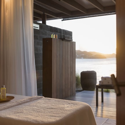 A room with a bed, a wooden table with a bottle and a towel, a stone wall, and a view of the sea.
