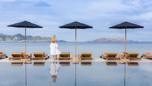 A poolside area with lounge chairs and umbrellas, a person in a white robe, and a view of the sea and hills in the background. - Christopher Hotel (St. Barthelemy)