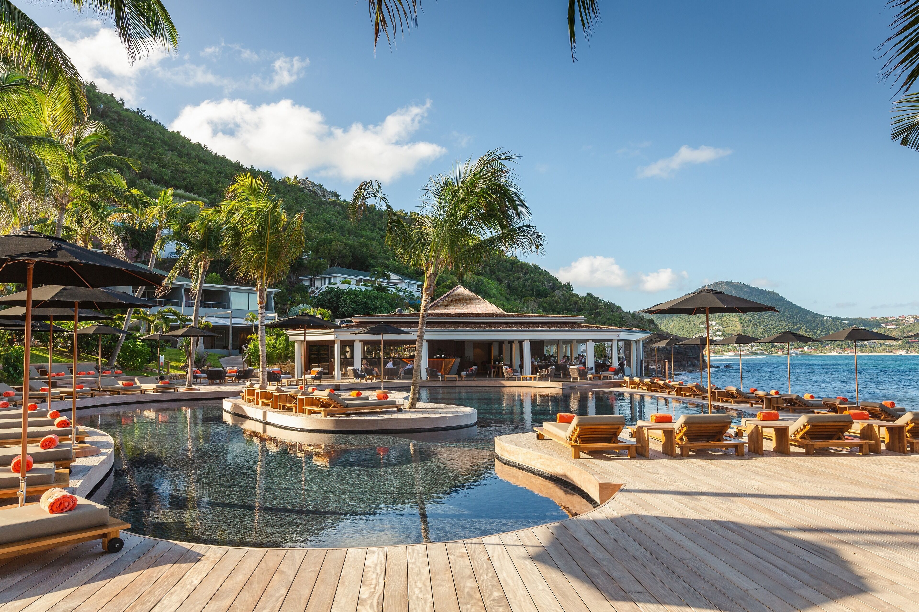 A resort pool area with lounge chairs and umbrellas, surrounded by palm trees and a view of the sea.