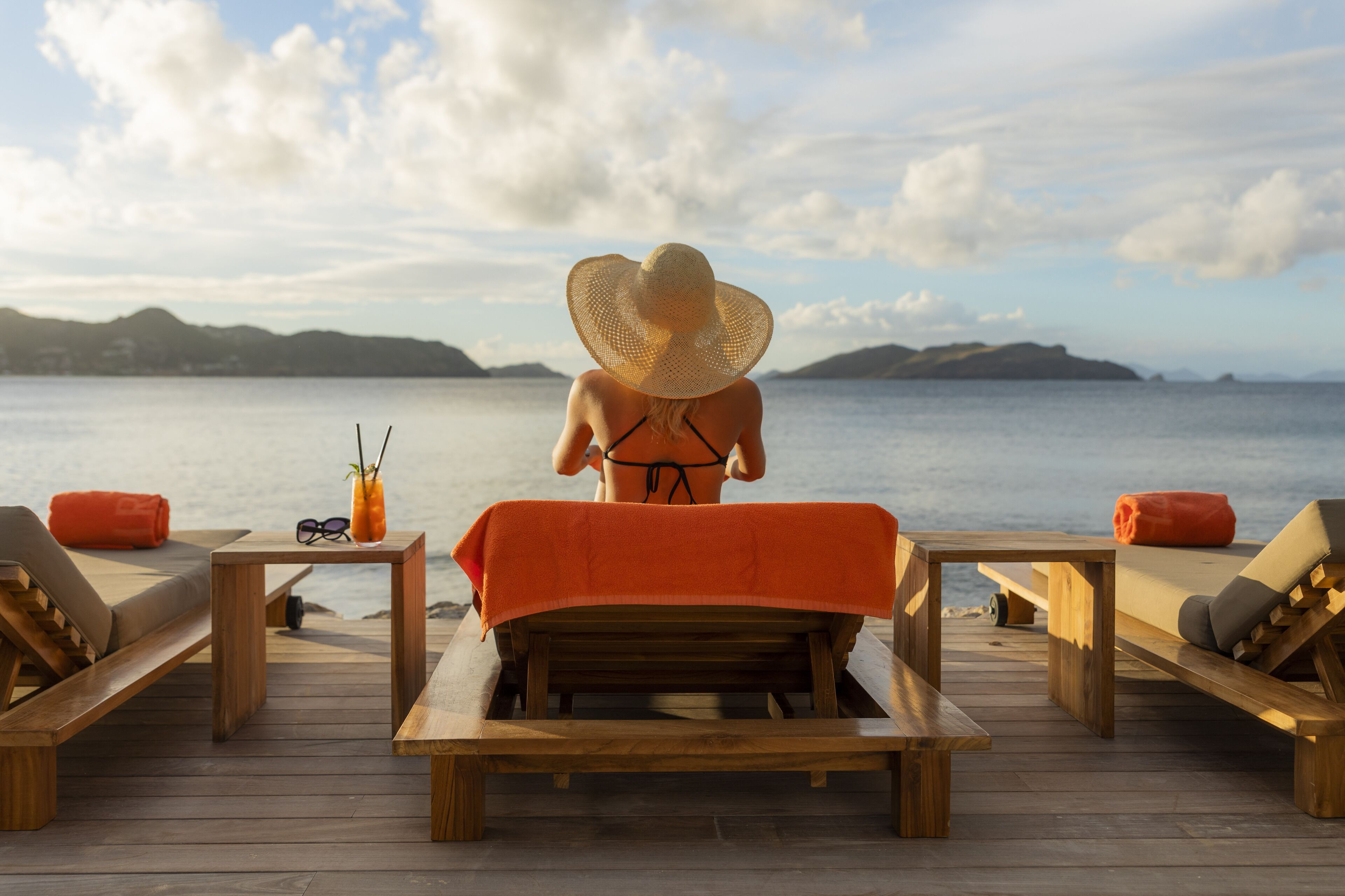 A wooden deck with lounge chairs and a table, overlooking a calm sea and distant hills.