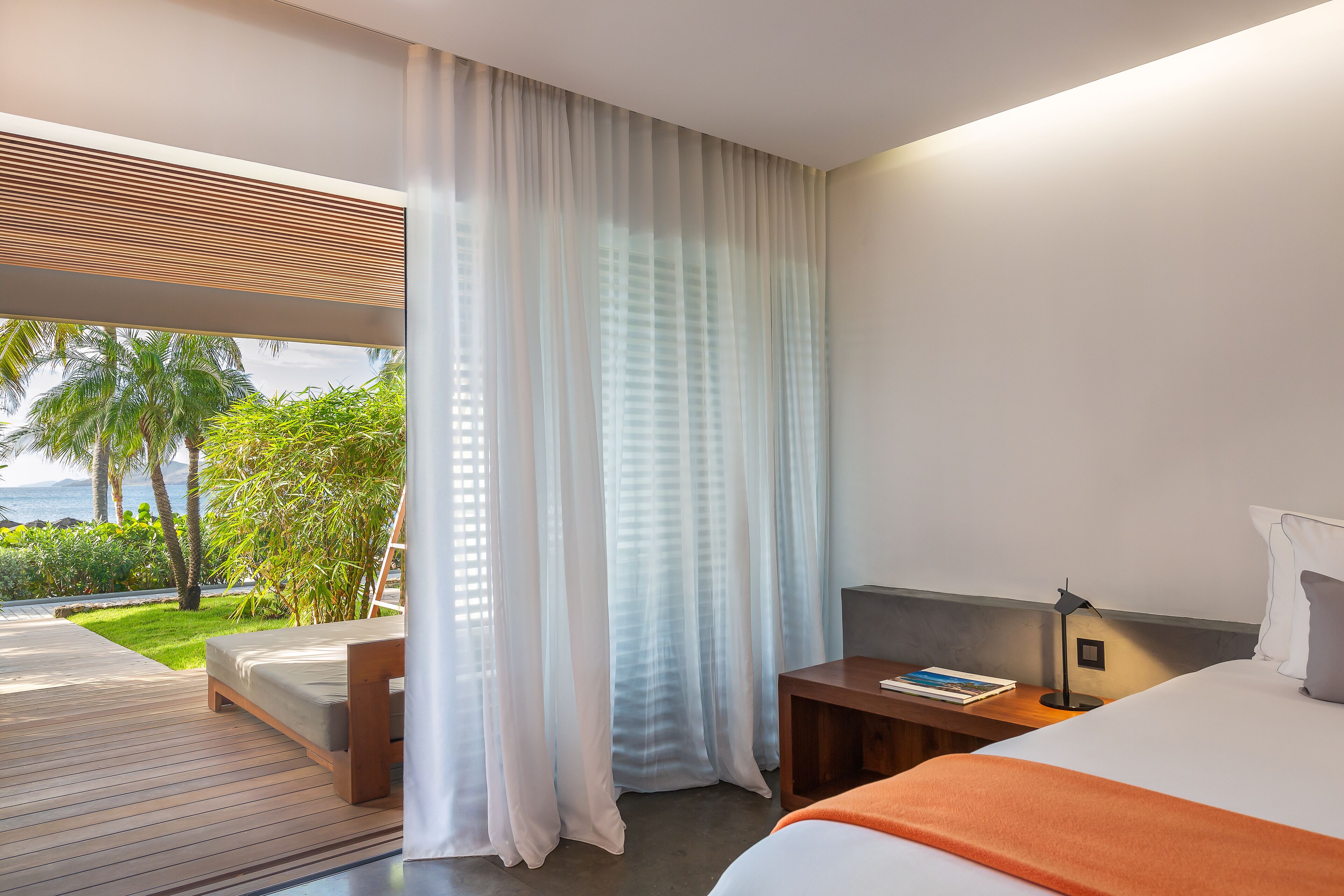 Une chambre d’hôtel moderne avec un balcon, un lit, une table de chevet avec une lampe, et une vue sur les palmiers et la mer.