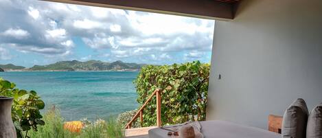 A bedroom with a view of the sea, a wooden deck, and a large potted plant.