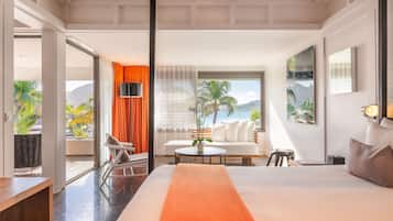 A modern hotel room with a large bed, a sofa, a dining table, and a balcony with a view of palm trees and mountains.