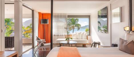 Habitación de hotel moderna con una cama grande, un sofá, una mesa de comedor y un balcón con vistas a palmeras y montañas.