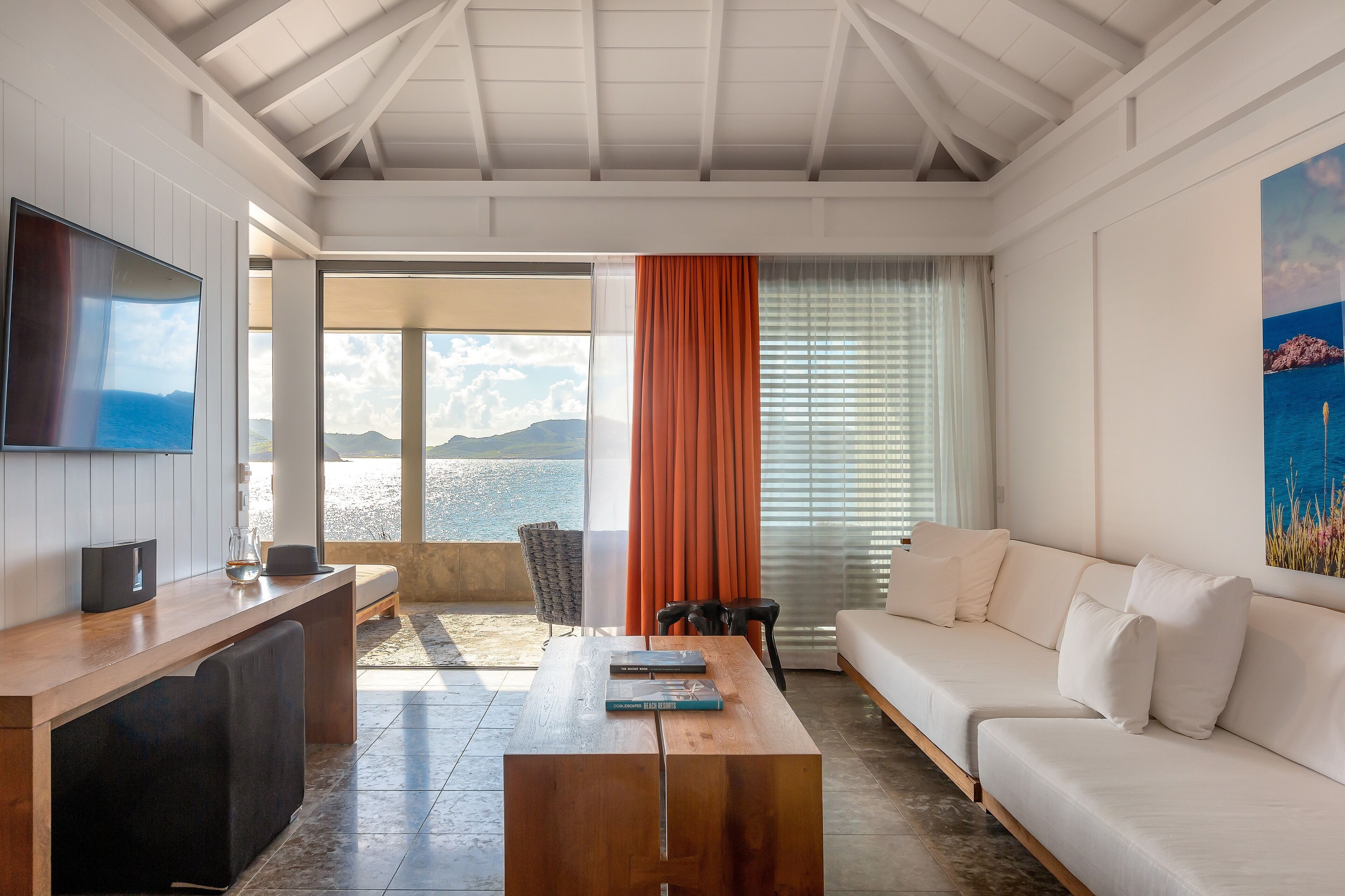 Una sala de estar moderna con una mesa de centro de madera, un sofá blanco, un televisor de pantalla plana y un amplio ventanal con vista al mar.