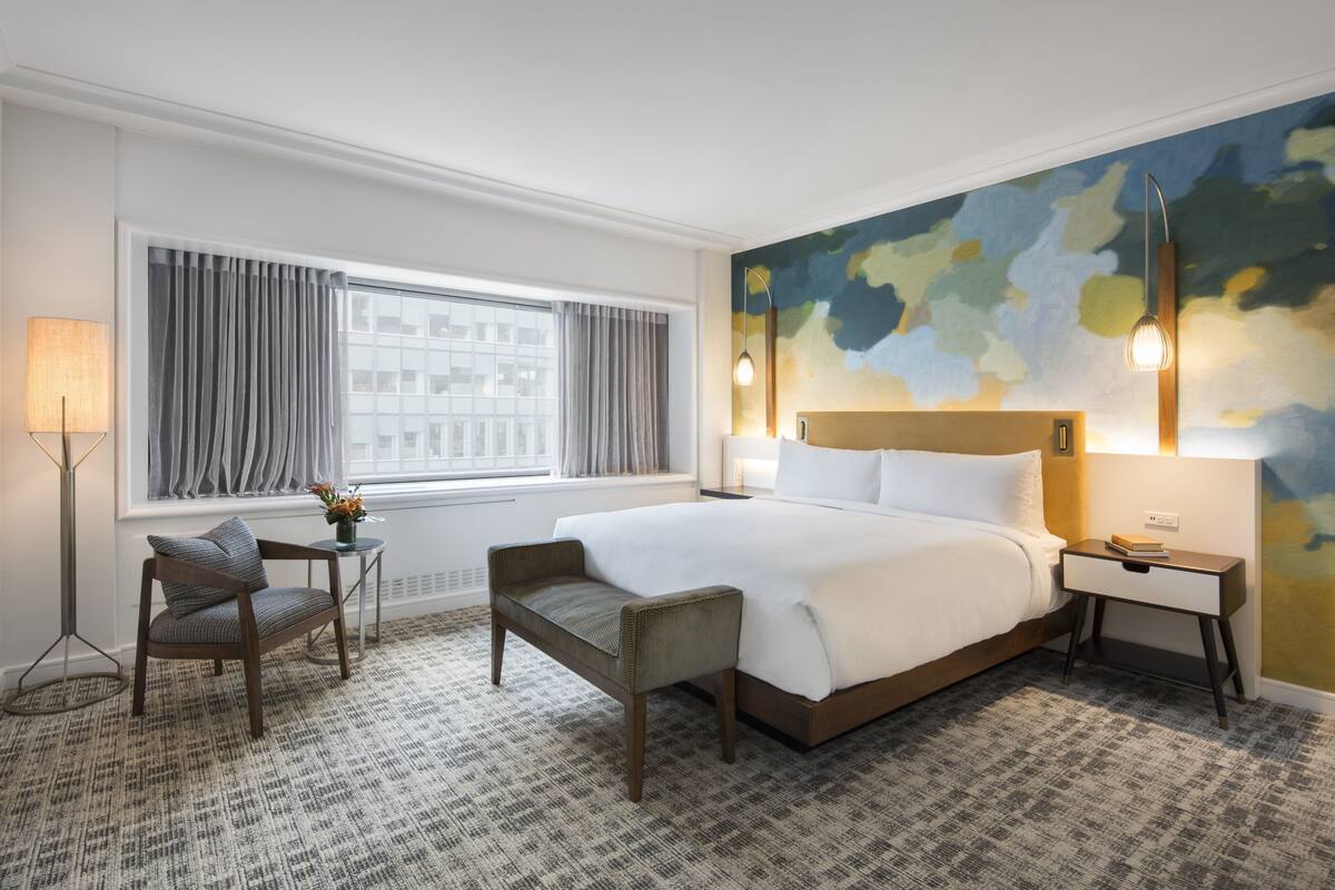 A hotel room with a large bed, a chair, a small table, a lamp, and a painting on the wall.