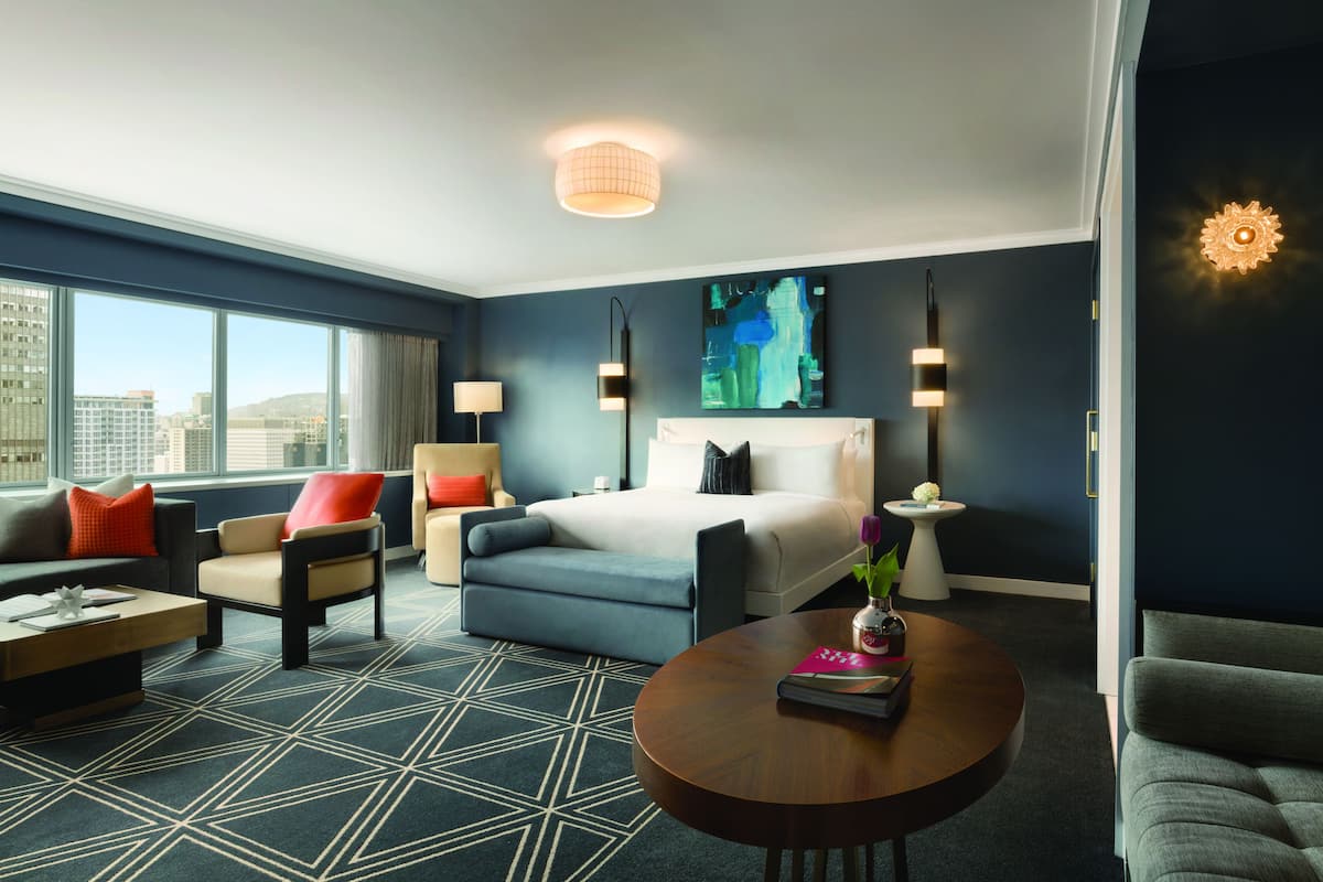 A modern hotel room with a large bed, a sofa, a chair, a coffee table, and a view of the city through large windows.