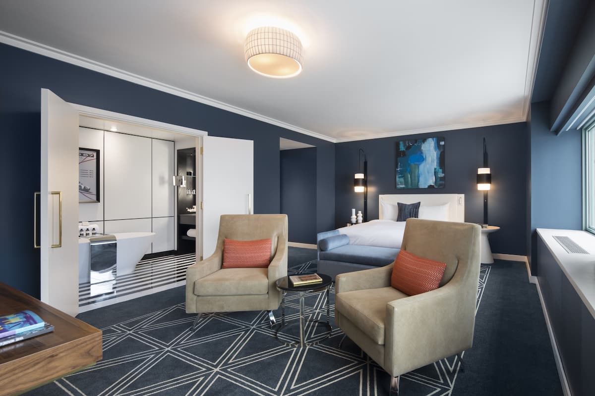 A modern hotel room with a bed, two armchairs, a coffee table, a desk, and a large window.