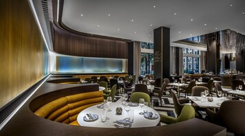A modern restaurant interior with round tables, comfortable seating, and a large window offering a view of the city.