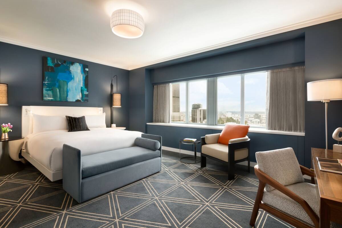 A modern hotel room with a large bed, a seating area with a chair and table, a view of the city through large windows, and a geometric patterned carpet.