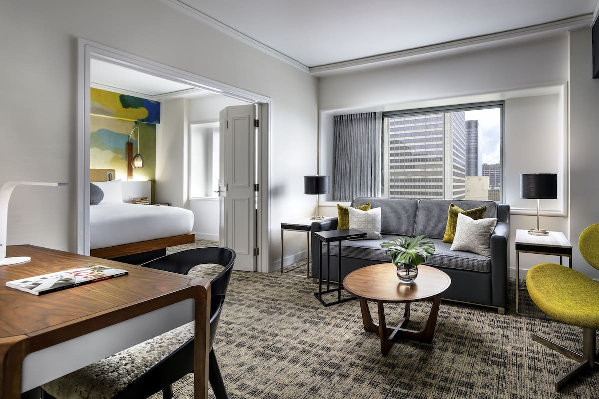 A modern hotel room with a bed, a sofa, a dining table, and a small coffee table. There is a window with a view of buildings.