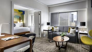 A modern hotel room with a bed, a sofa, a dining table, and a small coffee table. There is a window with a view of buildings.