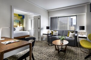 Habitación de hotel moderna con cama, sofá, mesa de comedor y una mesita de centro. Se ve una ventana con vistas a edificios.