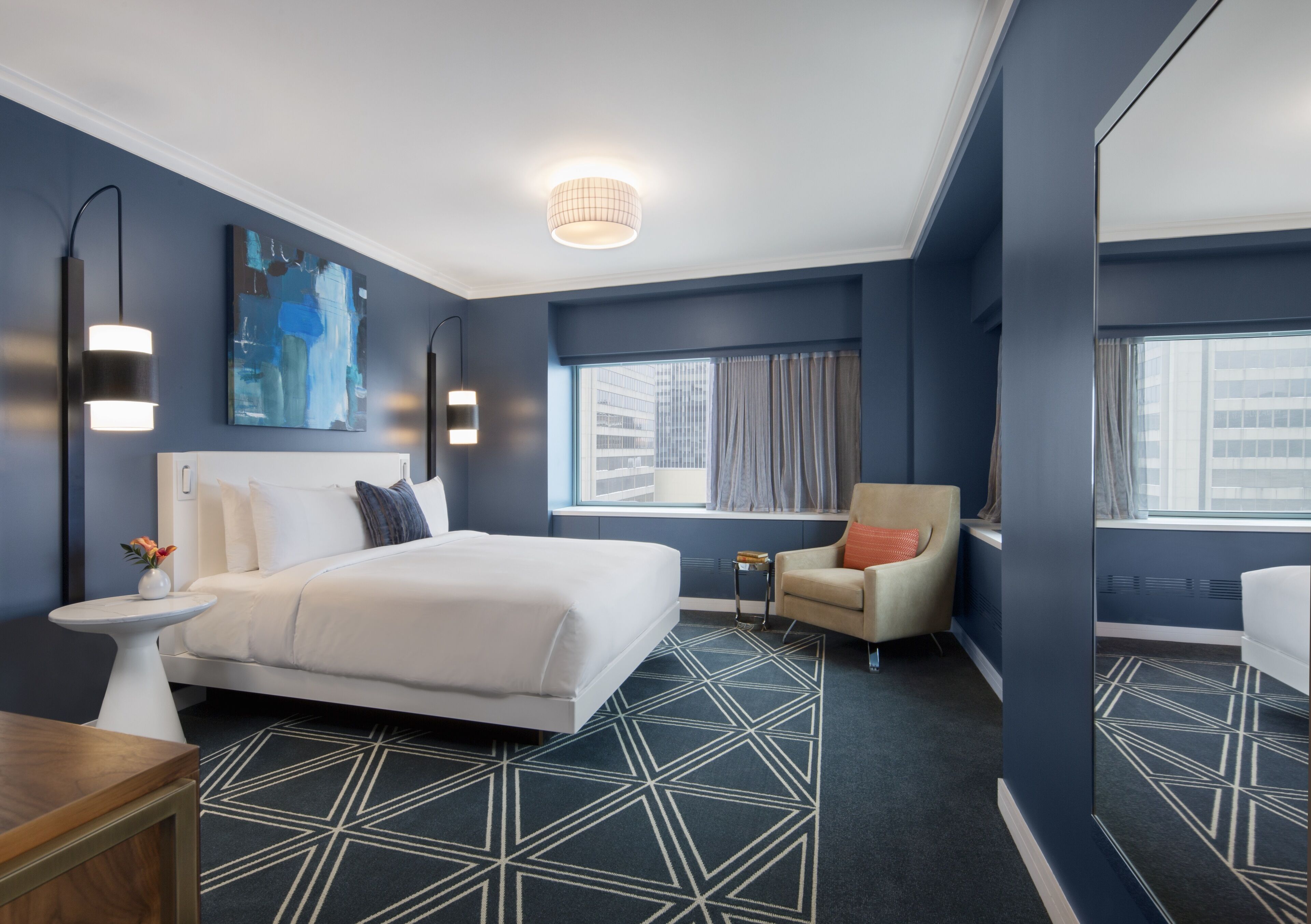 A modern hotel room with a large bed, a small table, a chair, and a large window with curtains.