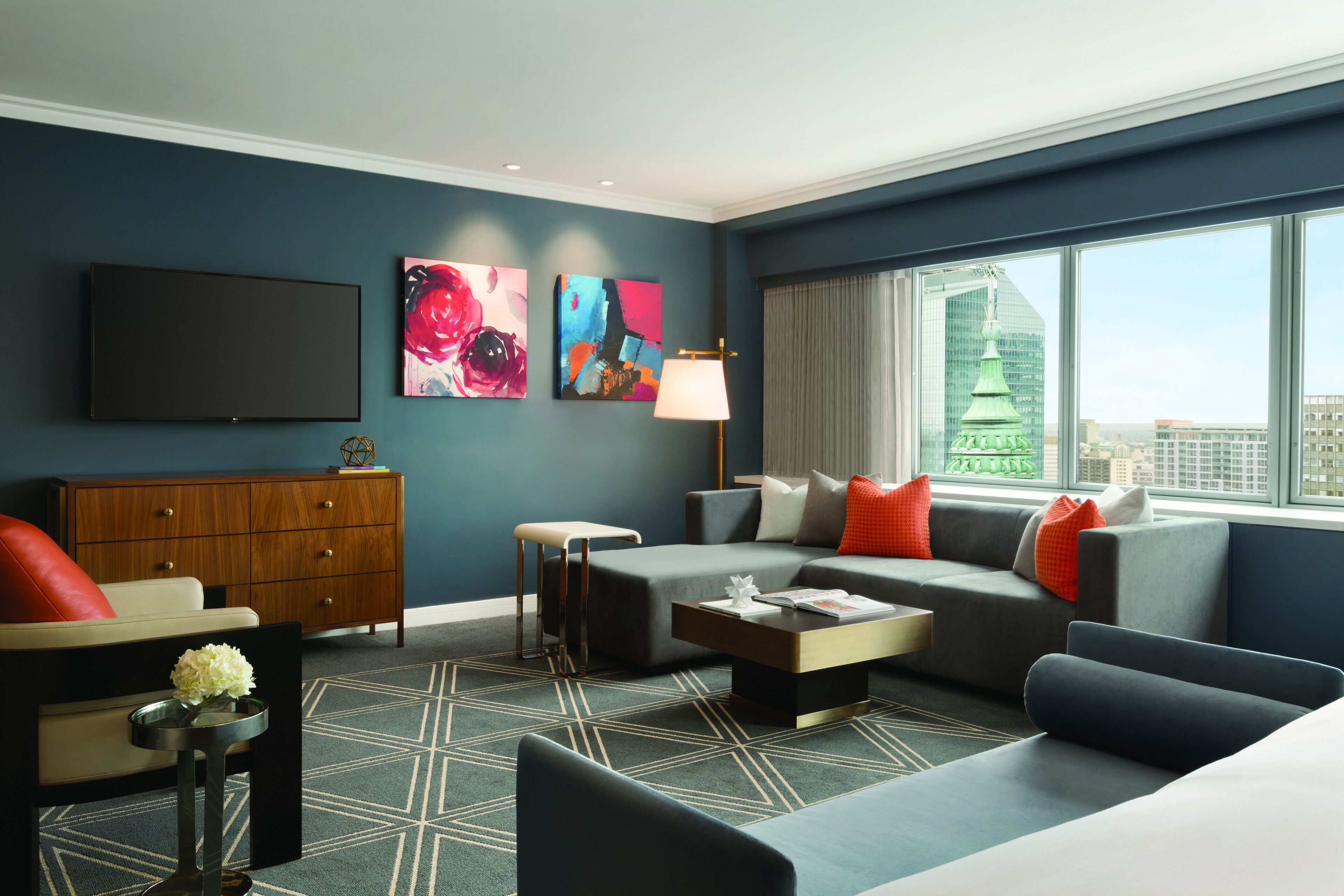 A modern hotel room with a large window, a flat-screen TV, a sofa, a coffee table, and artwork on the walls.