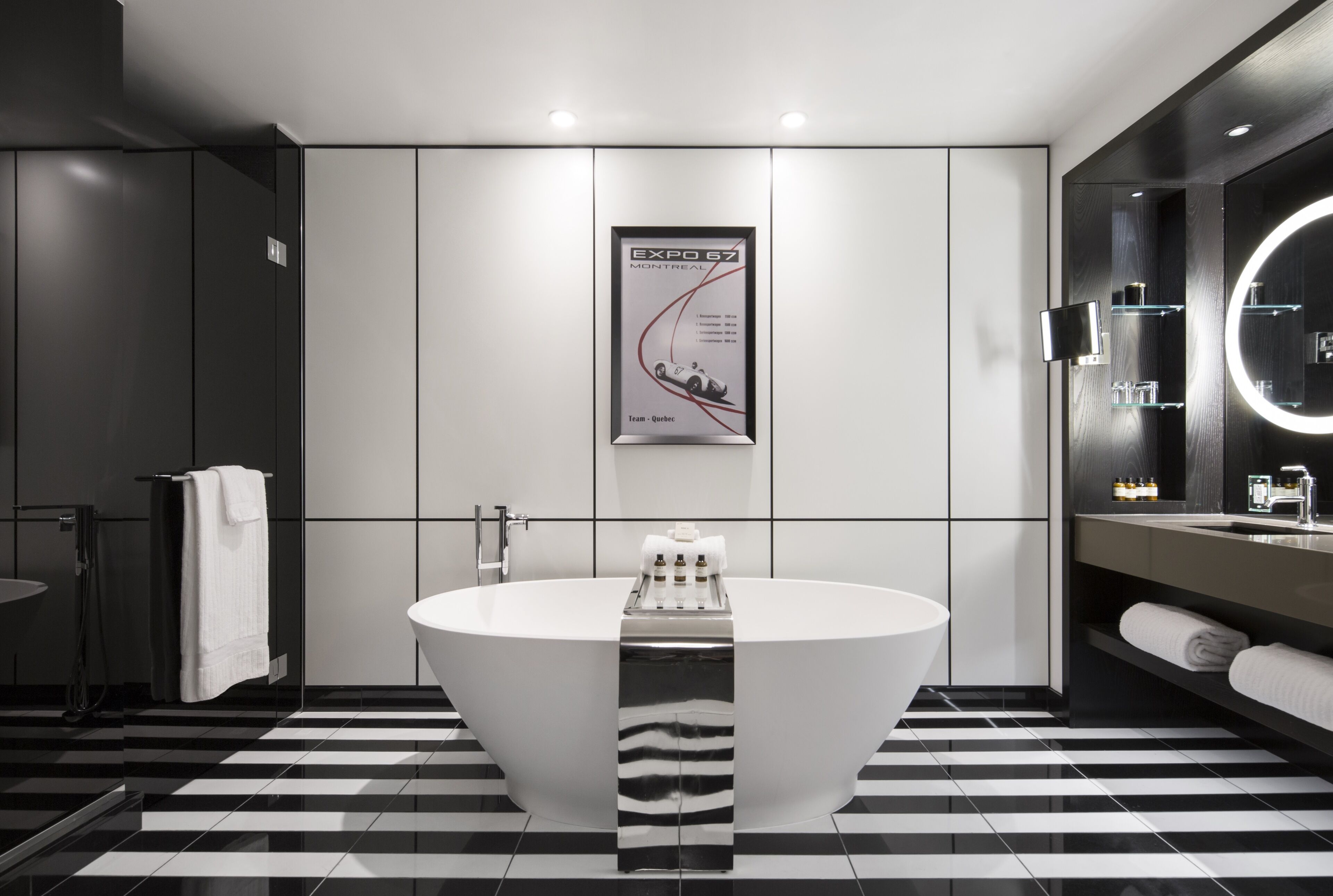 A modern bathroom with a large freestanding bathtub, a mirror with a circular light, and a poster on the wall.