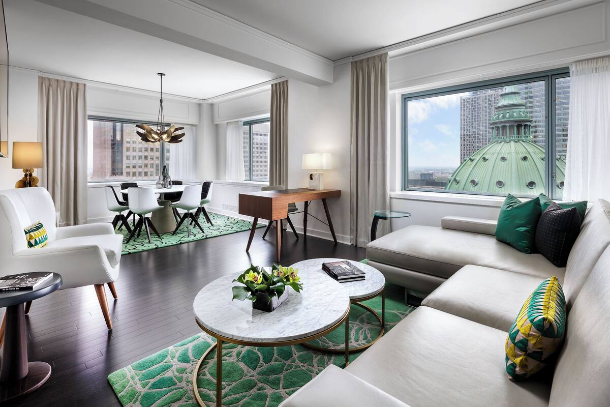A modern hotel room with a dining area, a sofa with decorative pillows, a round coffee table, and a view of a cityscape through large windows.
