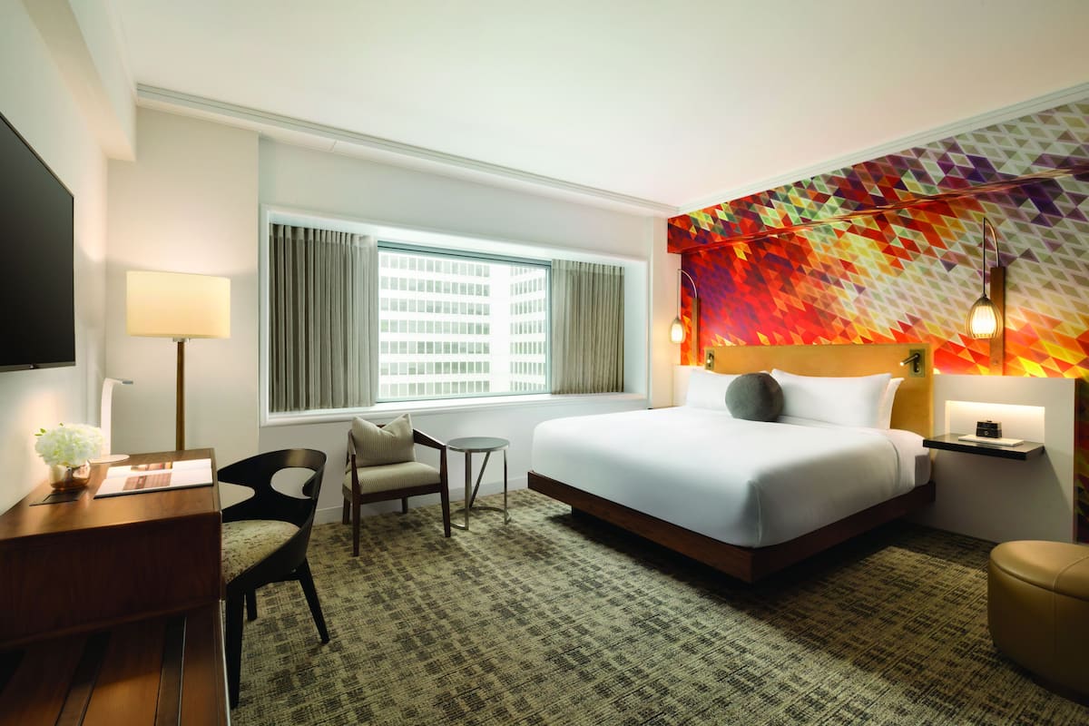 A hotel room with a large bed, a desk with a lamp, a chair, and a view of a building through the window.