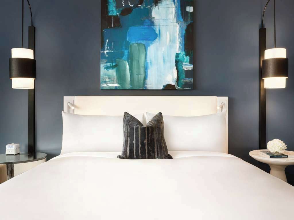 Une chambre d’hôtel moderne avec un grand lit, une tête de lit blanche, deux lampes de chevet, une table de chevet avec une horloge et une œuvre d’art murale.