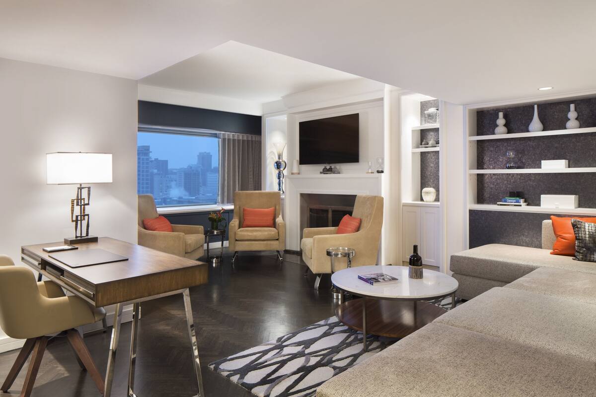 A modern living room with a sofa, armchair, and coffee table, a dining area with a table and chairs, a fireplace, and a TV mounted on the wall.