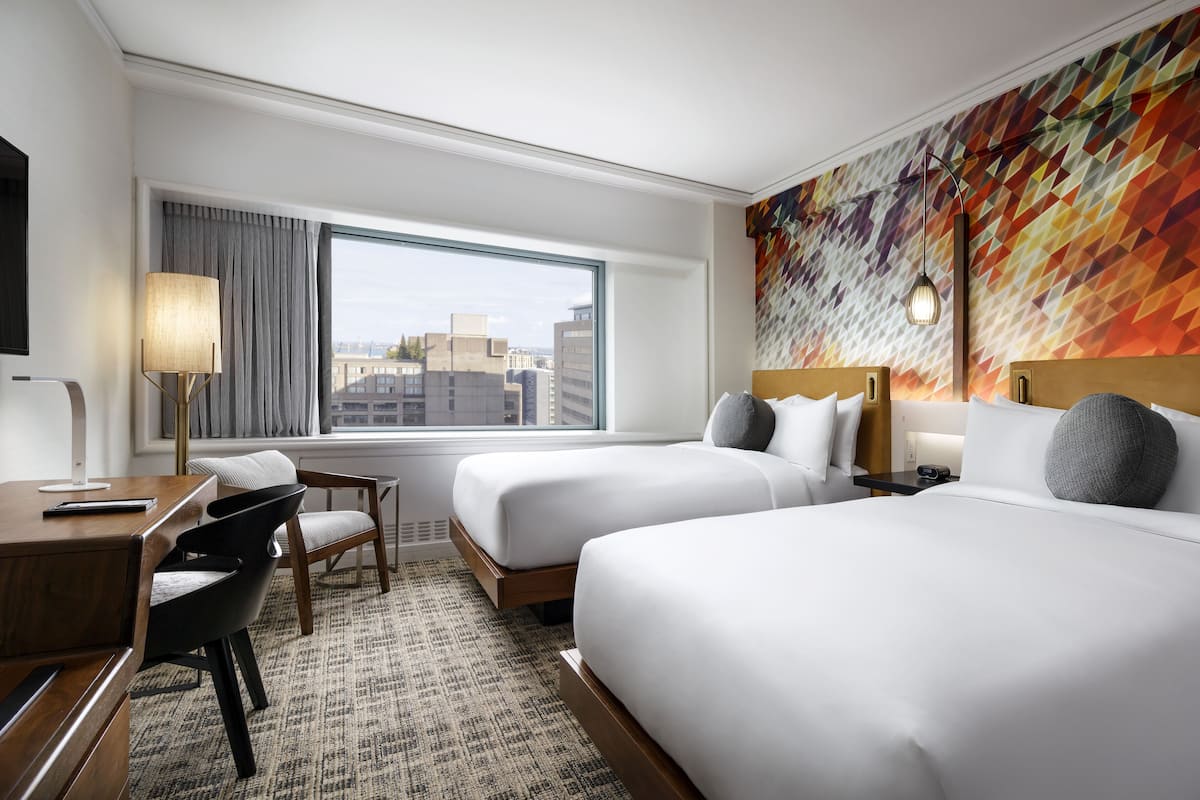 A hotel room with two beds, a desk with a chair, a lamp, and a view of the city through a large window.