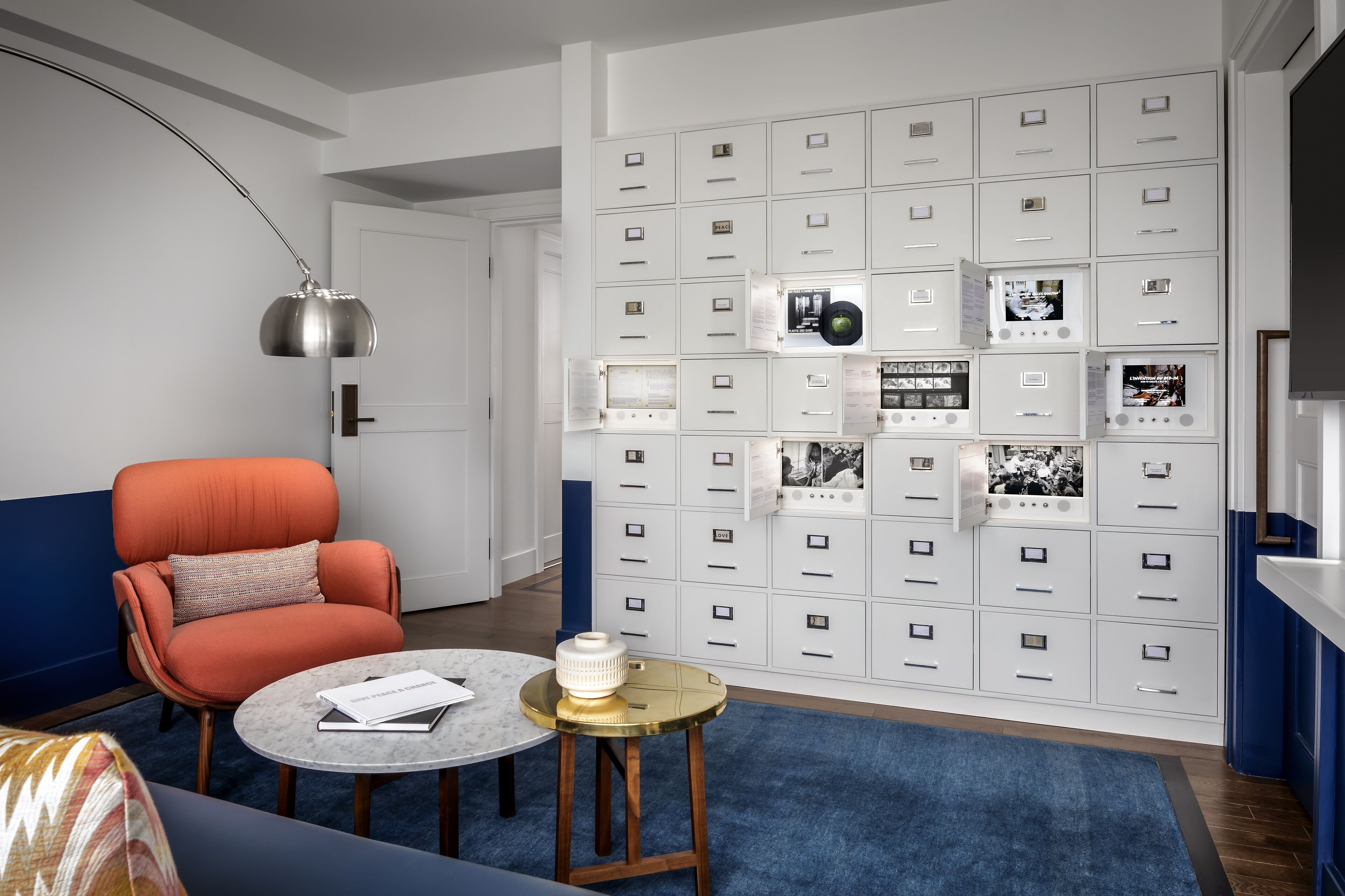 Uma sala de estar moderna com um sofá azul, uma poltrona laranja, uma mesa de centro redonda e uma parede de armários com fotos e documentos.