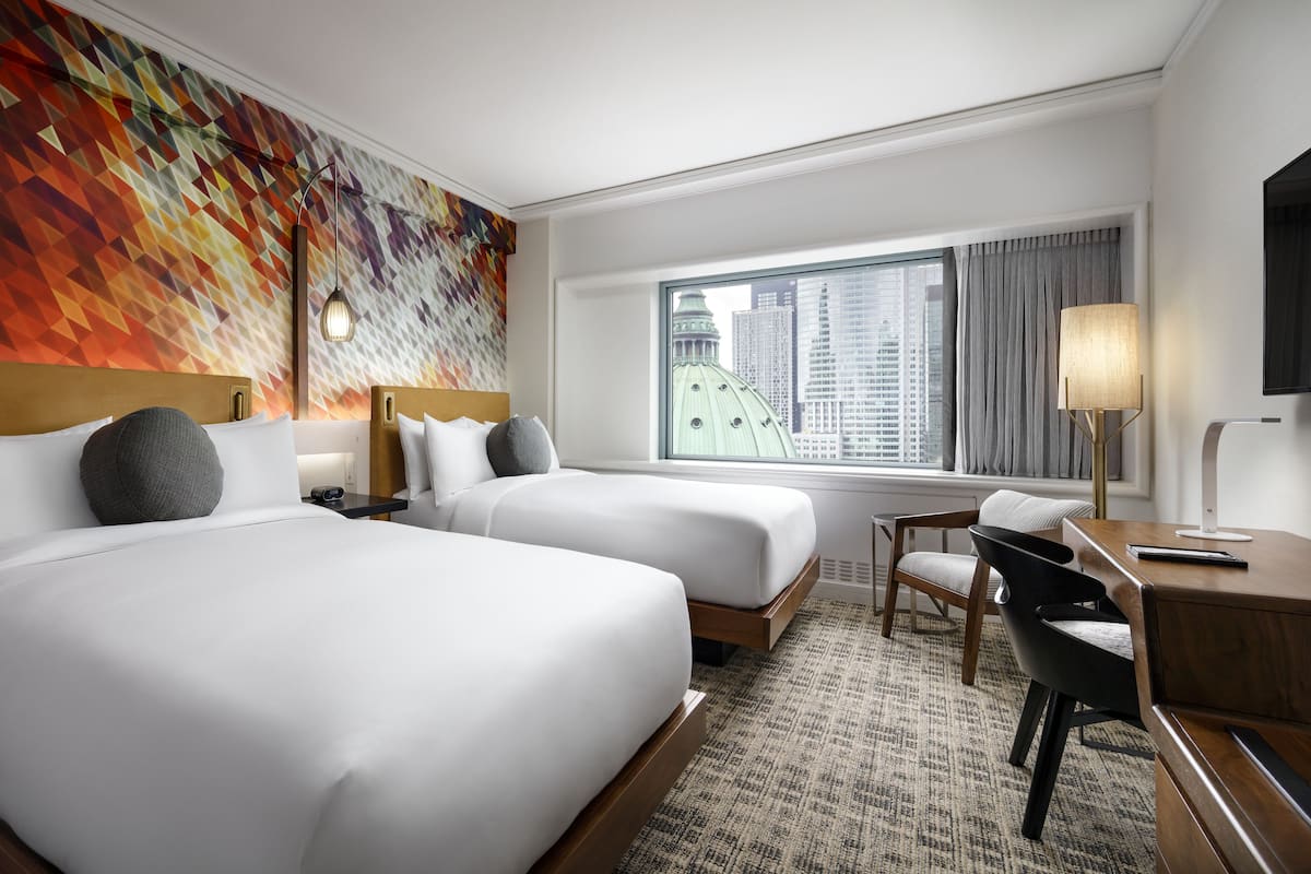 A hotel room with two beds, a desk, a chair, a lamp, and a large window with a city view.