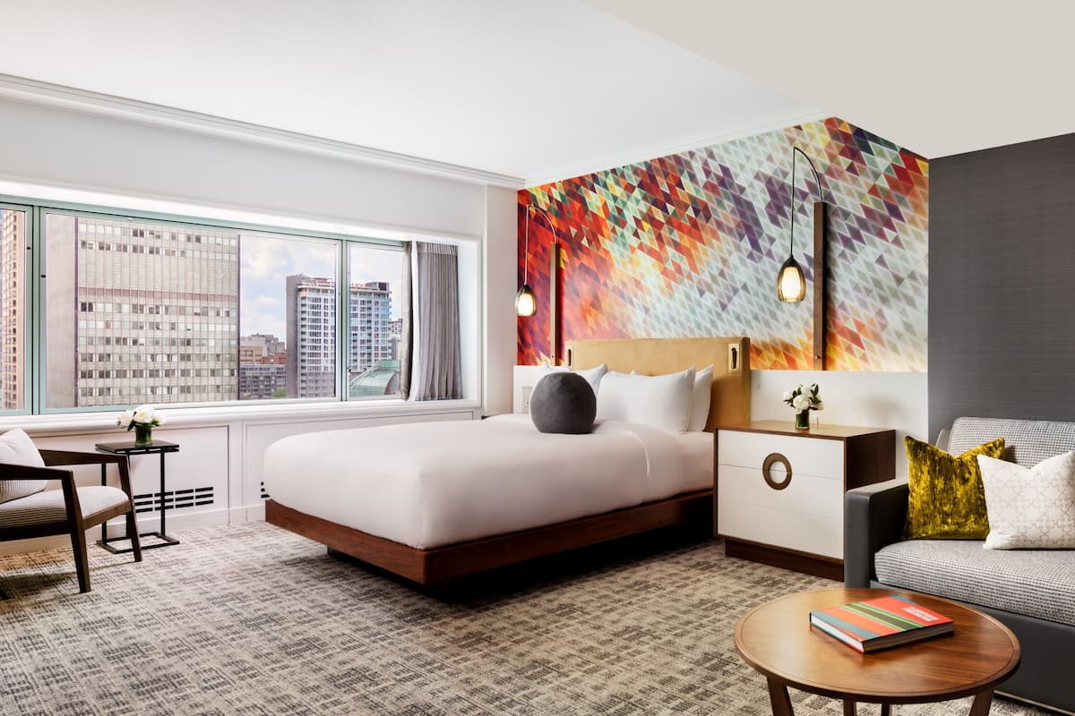 A modern hotel room with a large bed, a sofa, a small table, and a city view through the window.
