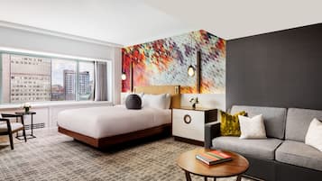 A modern hotel room with a large bed, a sofa, a small table, and a city view through the window.