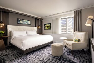 A hotel room with a large bed, a small round table, a chair with a decorative pillow, and a large window with curtains.
