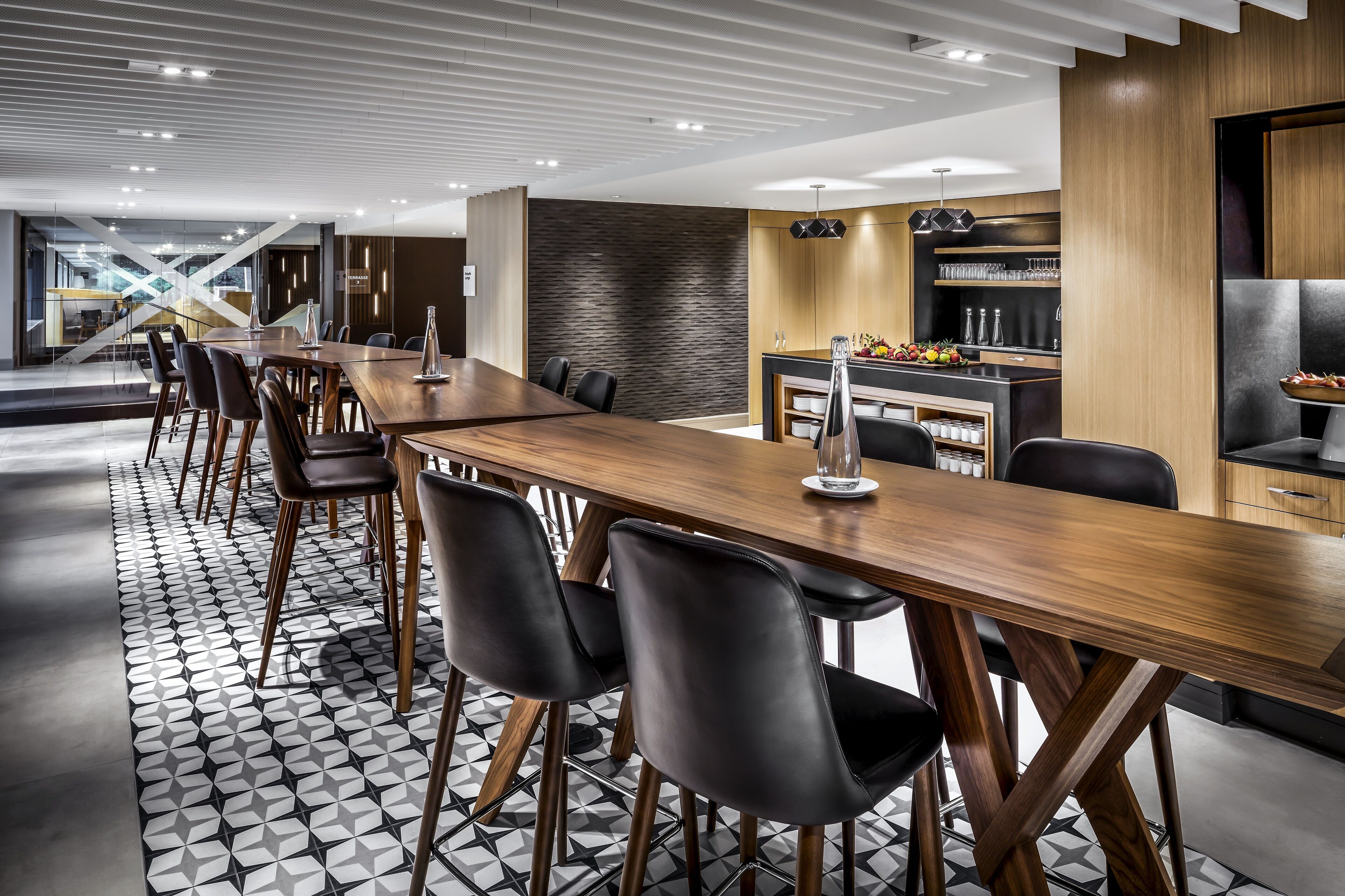 Un espace de salle à manger moderne avec une longue table en bois, des chaises noires et un sol orné d’un motif géométrique.