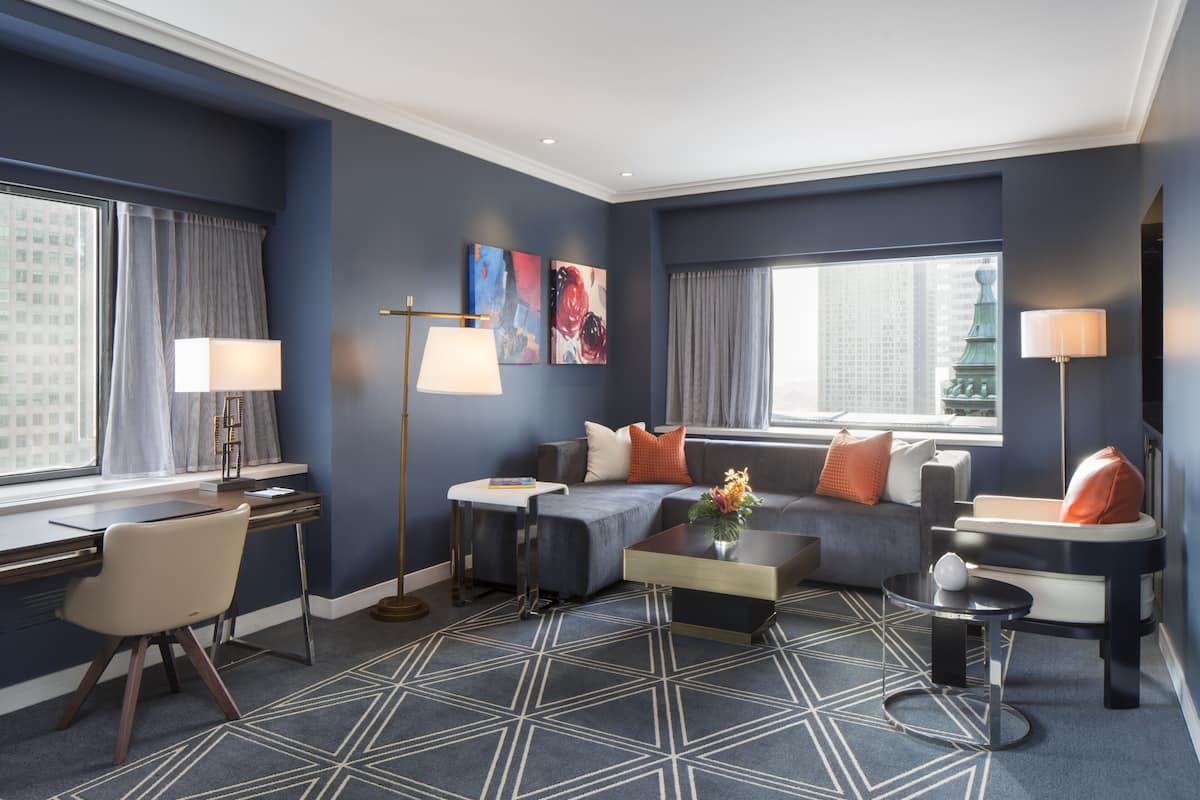 A modern hotel room with a dark blue accent wall, a large window with a view of the city, a sofa set, a desk with a chair, and a floor lamp.