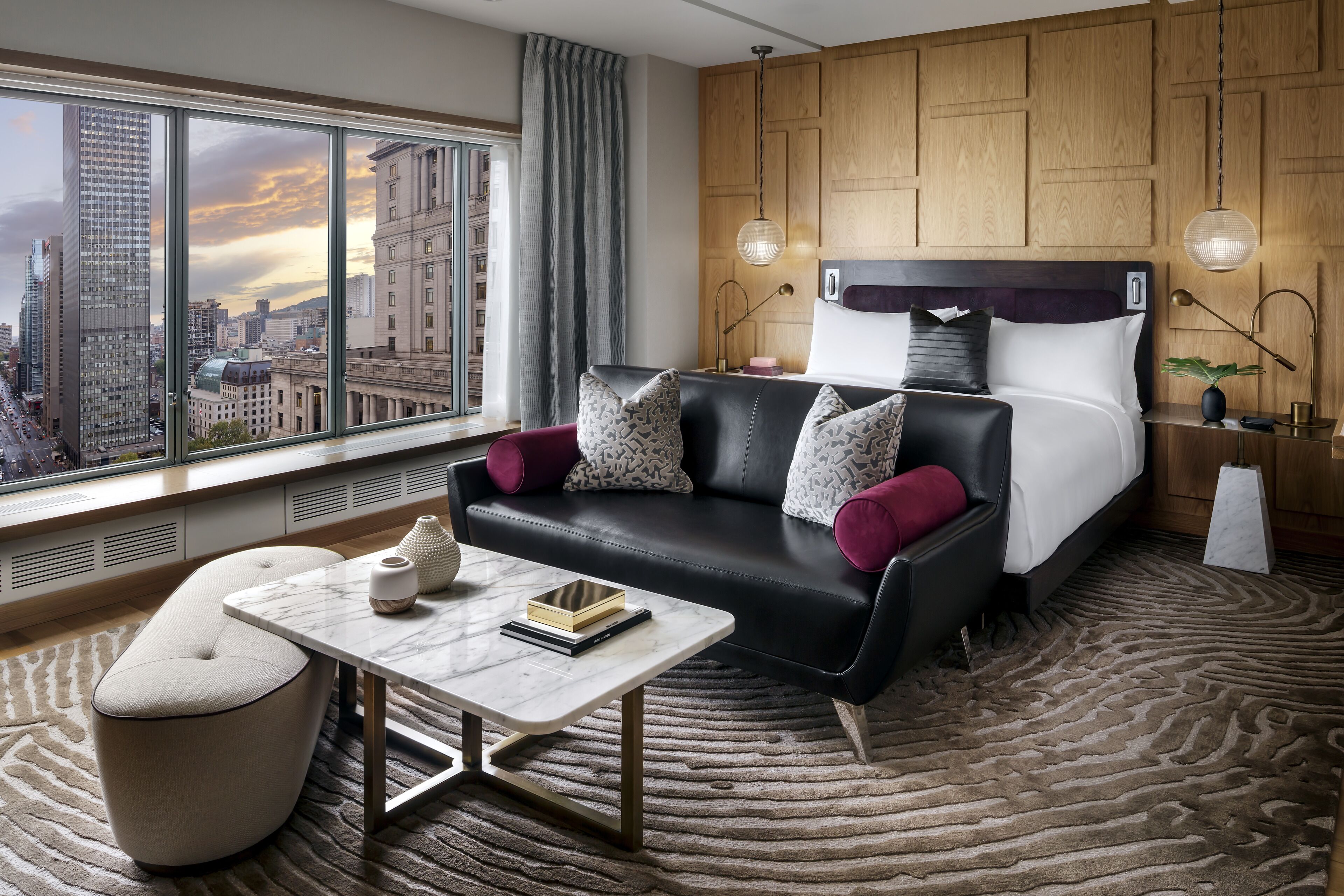 A modern hotel room with a large window, a black leather sofa, a marble coffee table, and a patterned rug.