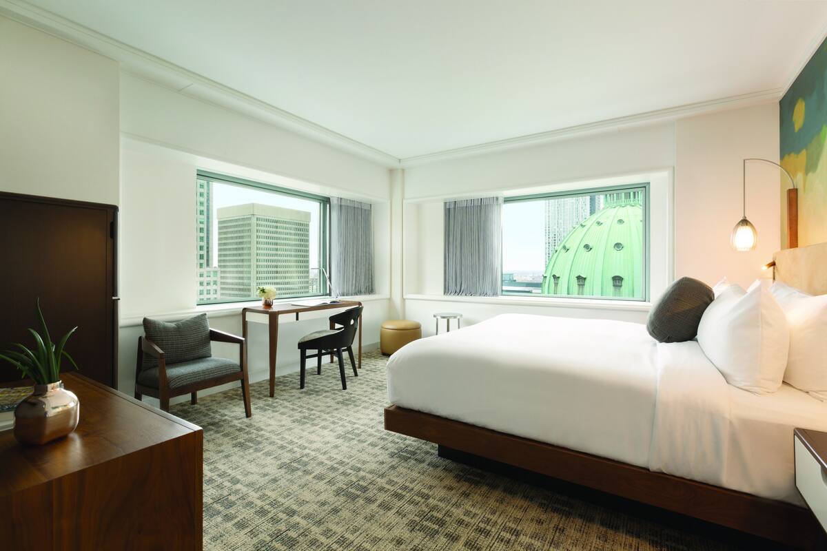 A hotel room with a large bed, a desk with a chair, a small table, and a potted plant.