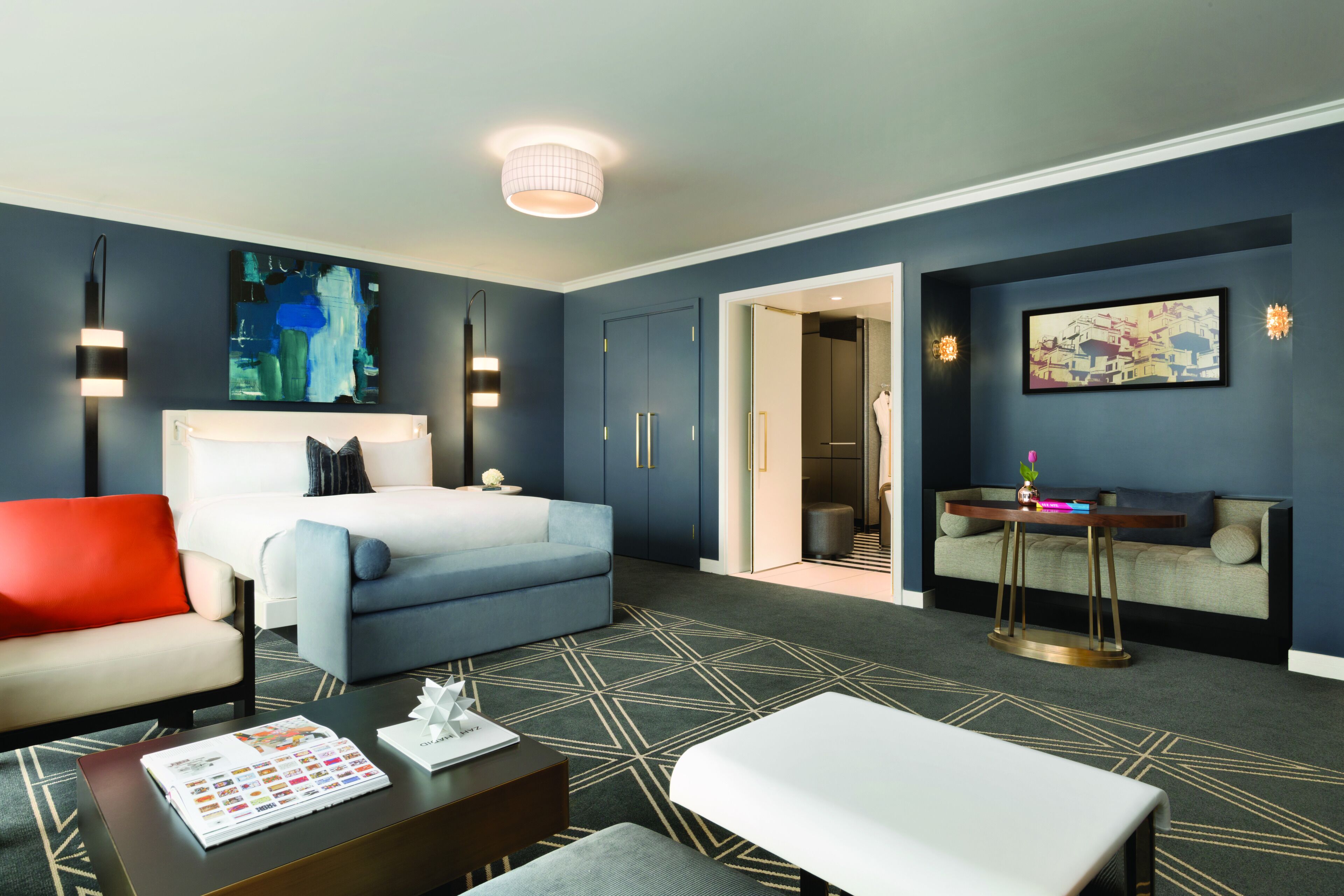 A modern hotel room with a bed, a sofa, a coffee table, and a desk. There are two doors, one leading to a bathroom and the other to a closet.