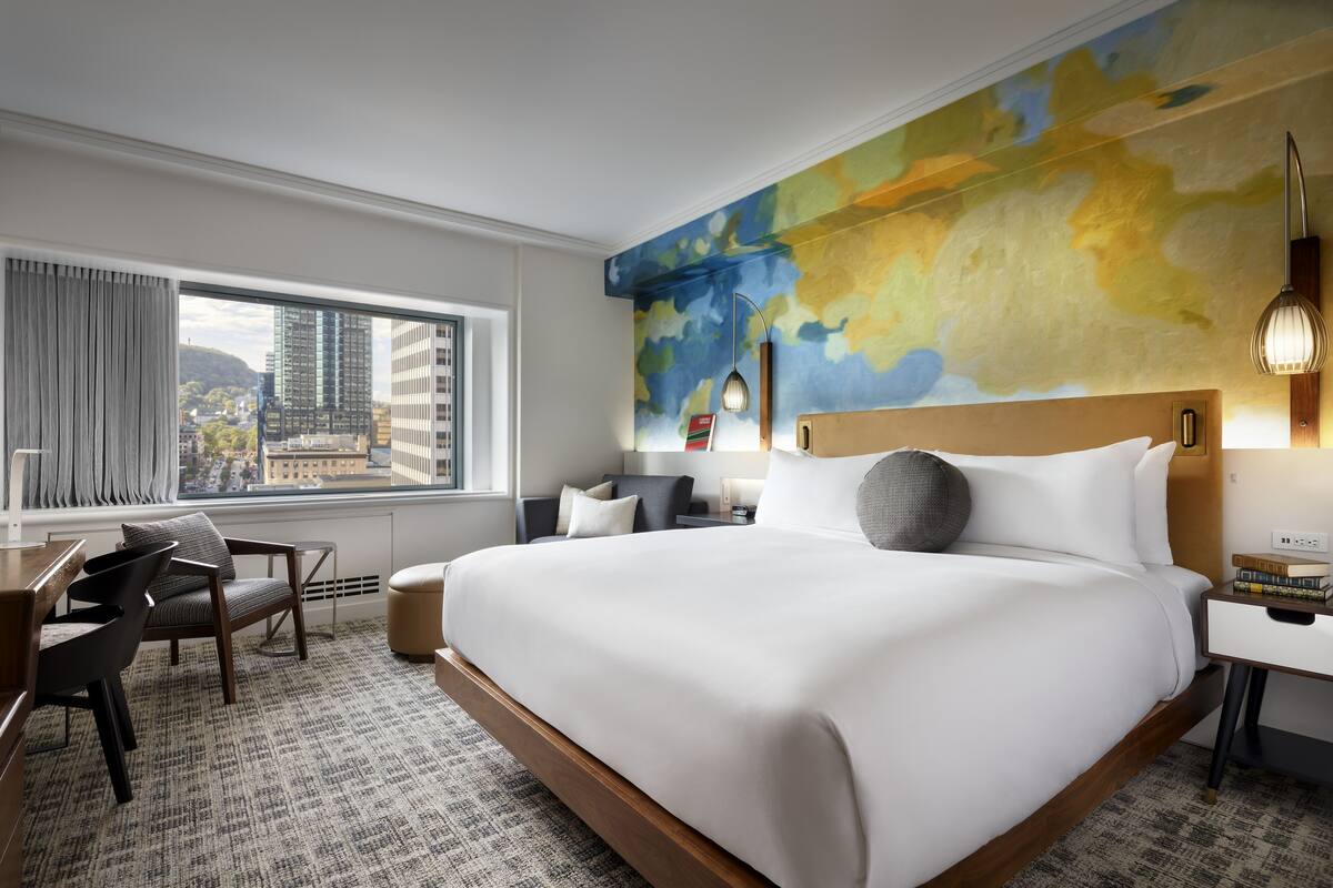 A hotel room with a large bed, a desk with a chair, a view of the city through the window, and a colorful abstract wall mural.