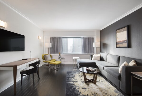 A modern hotel room with a flat-screen TV, a sofa, a yellow armchair, a desk, and a large window with curtains.