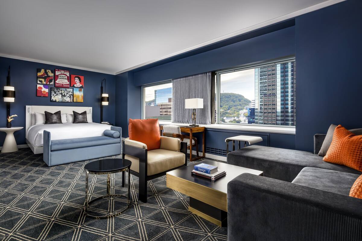 A modern hotel room with a bed, sofa, armchair, and a coffee table. There is a large window with a city view.