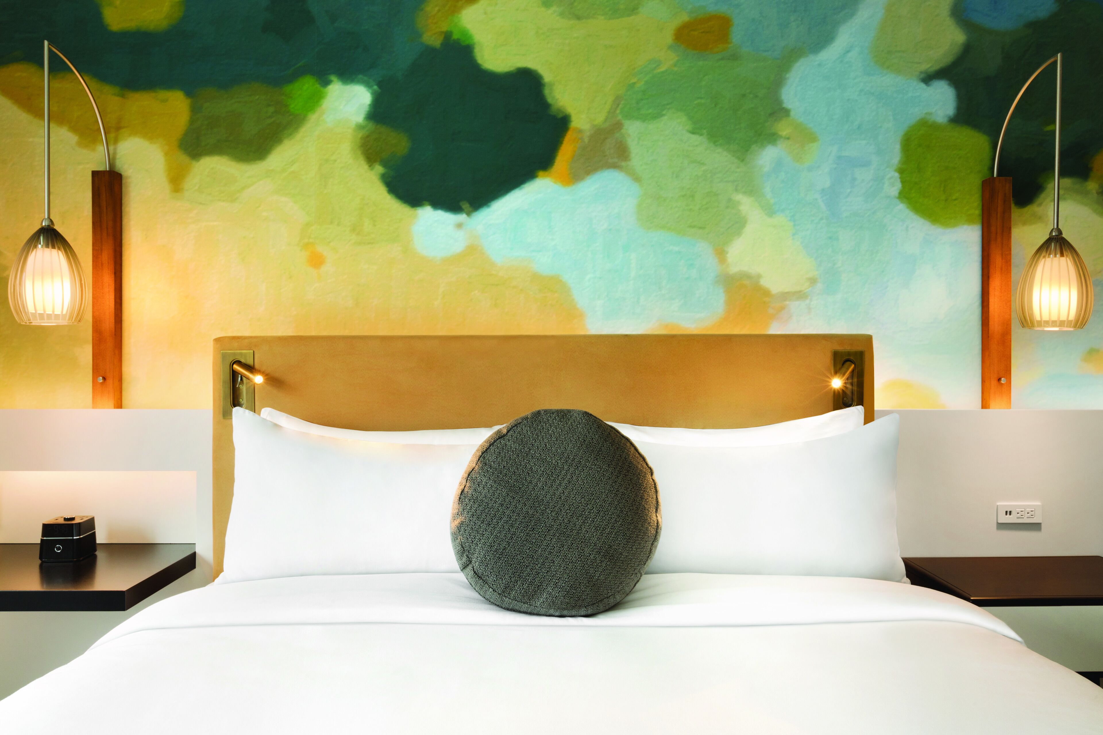 Una habitación de hotel con una cama moderna, un cojín redondo y un llamativo mural abstracto en la pared.