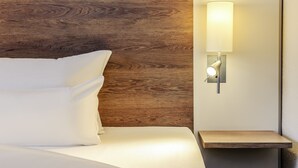 Una cama bien hecha con sábanas blancas, un cabecero de madera y una mesita de noche con una lámpara.