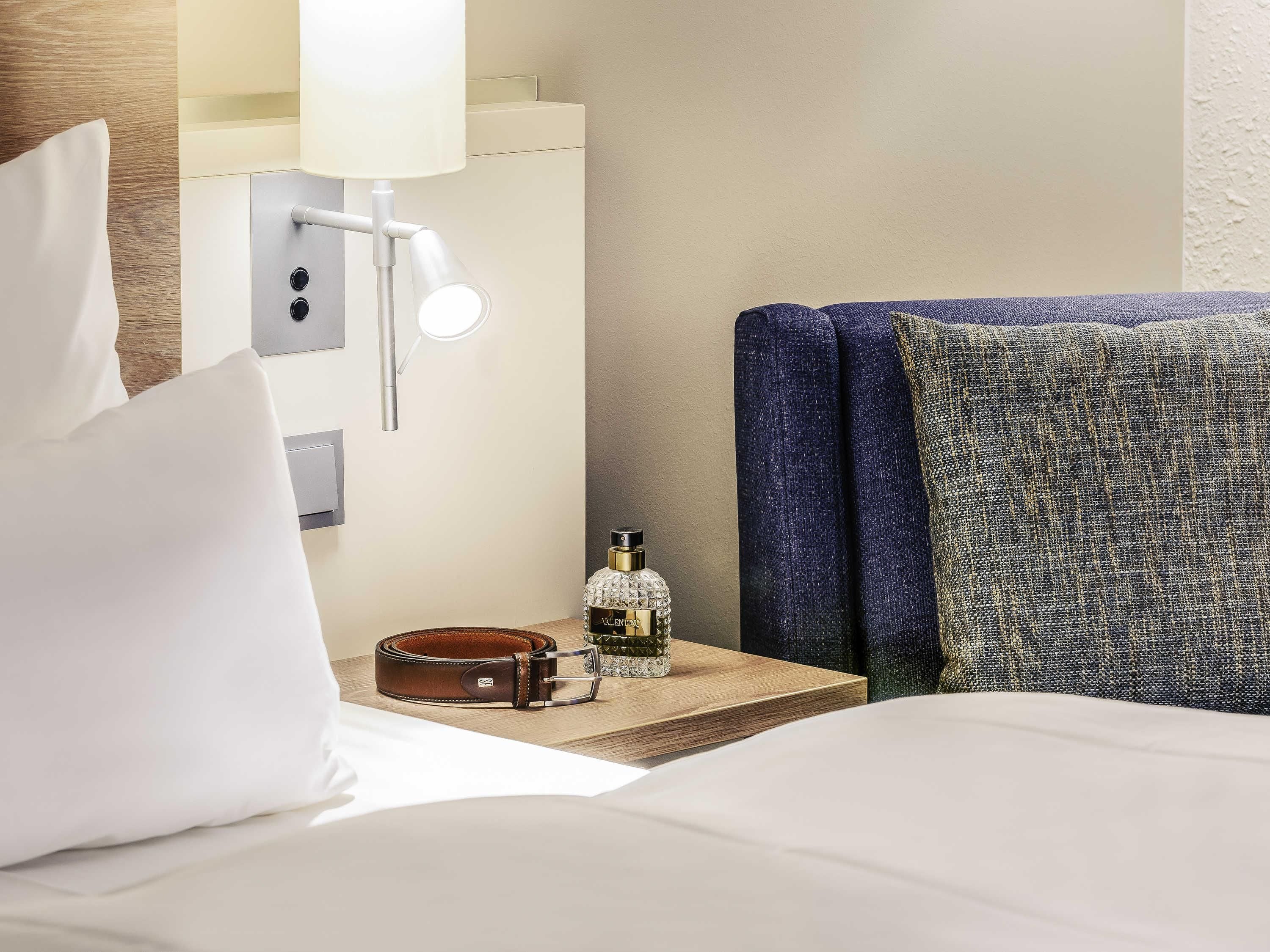 Una habitación de hotel con una cama, una mesita de noche, una lámpara, un frasco de perfume y un bolso de cuero marrón.